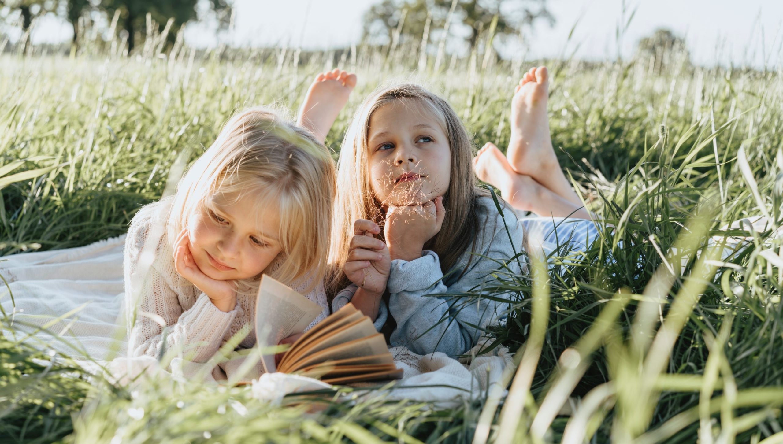 two young girls reading a book laying on the grass